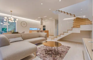 Bright modern living room with a sectional sofa, round coffee table, patterned rug, open wooden staircase, and minimalist lighting.