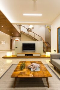 Modern living room with a wooden coffee table, wall-mounted TV unit, open staircase, warm lighting, and neutral furnishings.