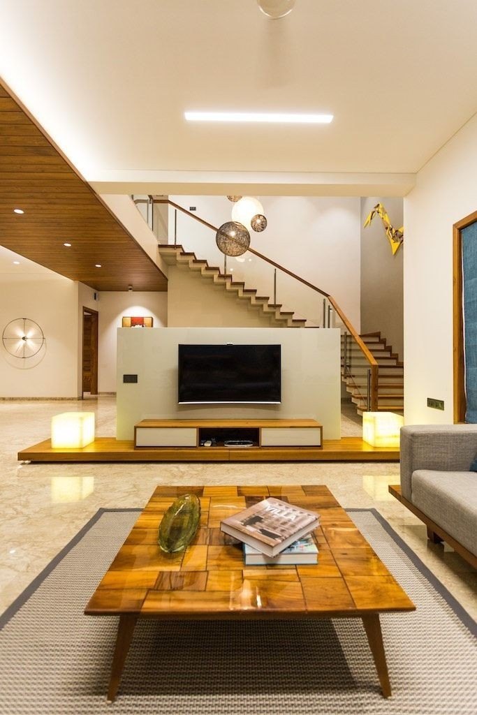 Modern living room with a wooden coffee table, wall-mounted TV unit, open staircase, warm lighting, and neutral furnishings.