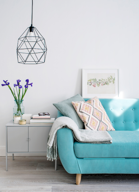 Modern living room interior featuring a teal sofa with decorative cushions, a minimalist side table with flowers, and a geometric pendant light against a white wall.